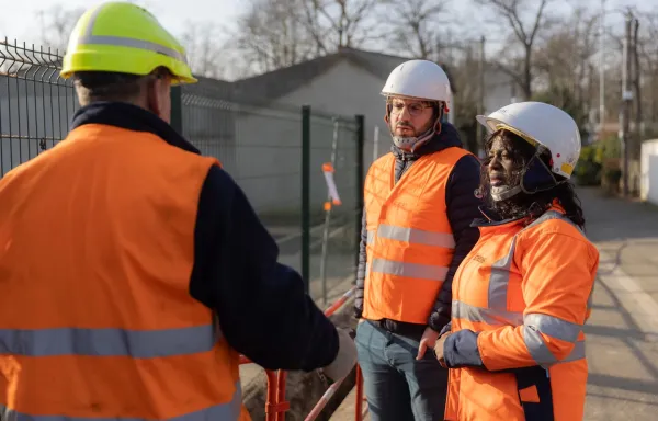 Échange entre techniciens en gilet orange sur un chantier urbain, portant des casques de sécurité