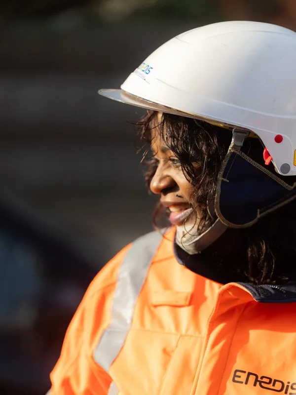 Technicienne Enedis souriante en tenue de sécurité orange et casque, sur un chantier