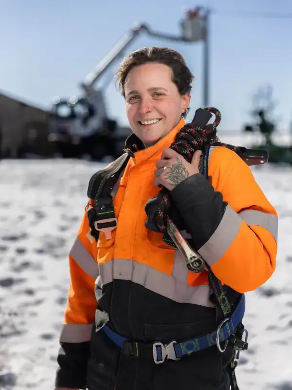 Monteuse de réseaux électriques souriante en tenue de sécurité, tenant du matériel de grimpe, sur un chantier enneigé